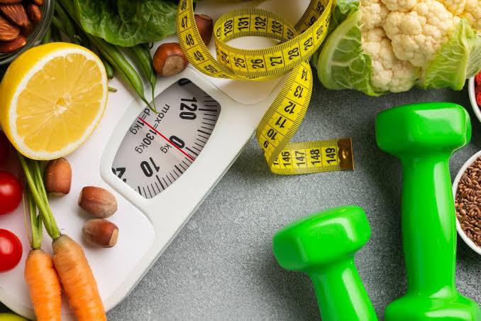 A scale, measuring tape, dumbbells, and fresh vegetables arranged on a stone surface