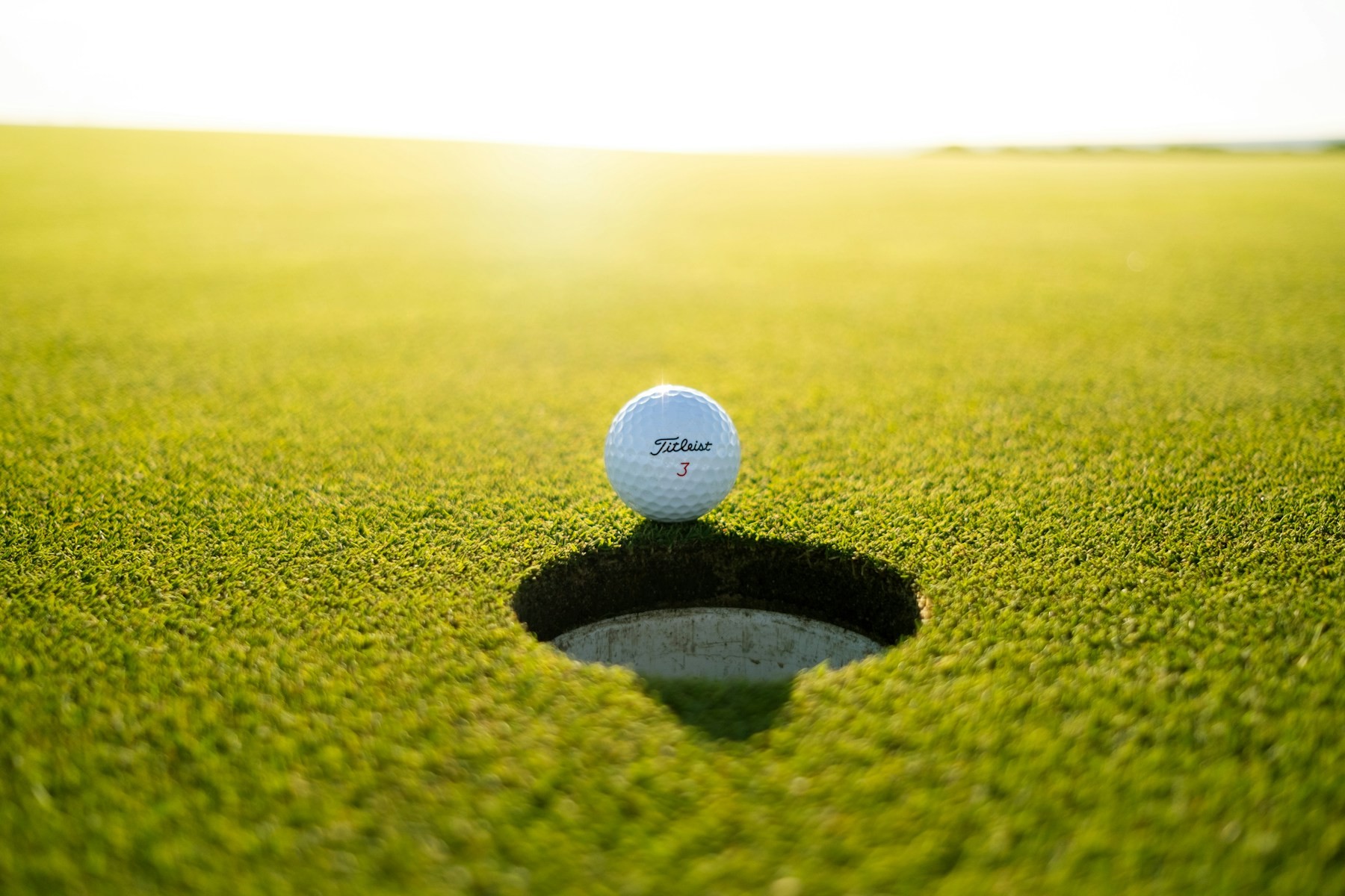 A golf ball resting at the edge of the hole on a sunlit green